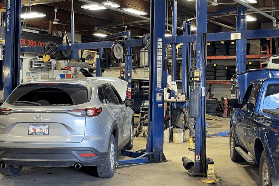 Garage auto repair bay with a silver SUV on a lift and a blue pickup nearby, tires stacked on shelves in the background.